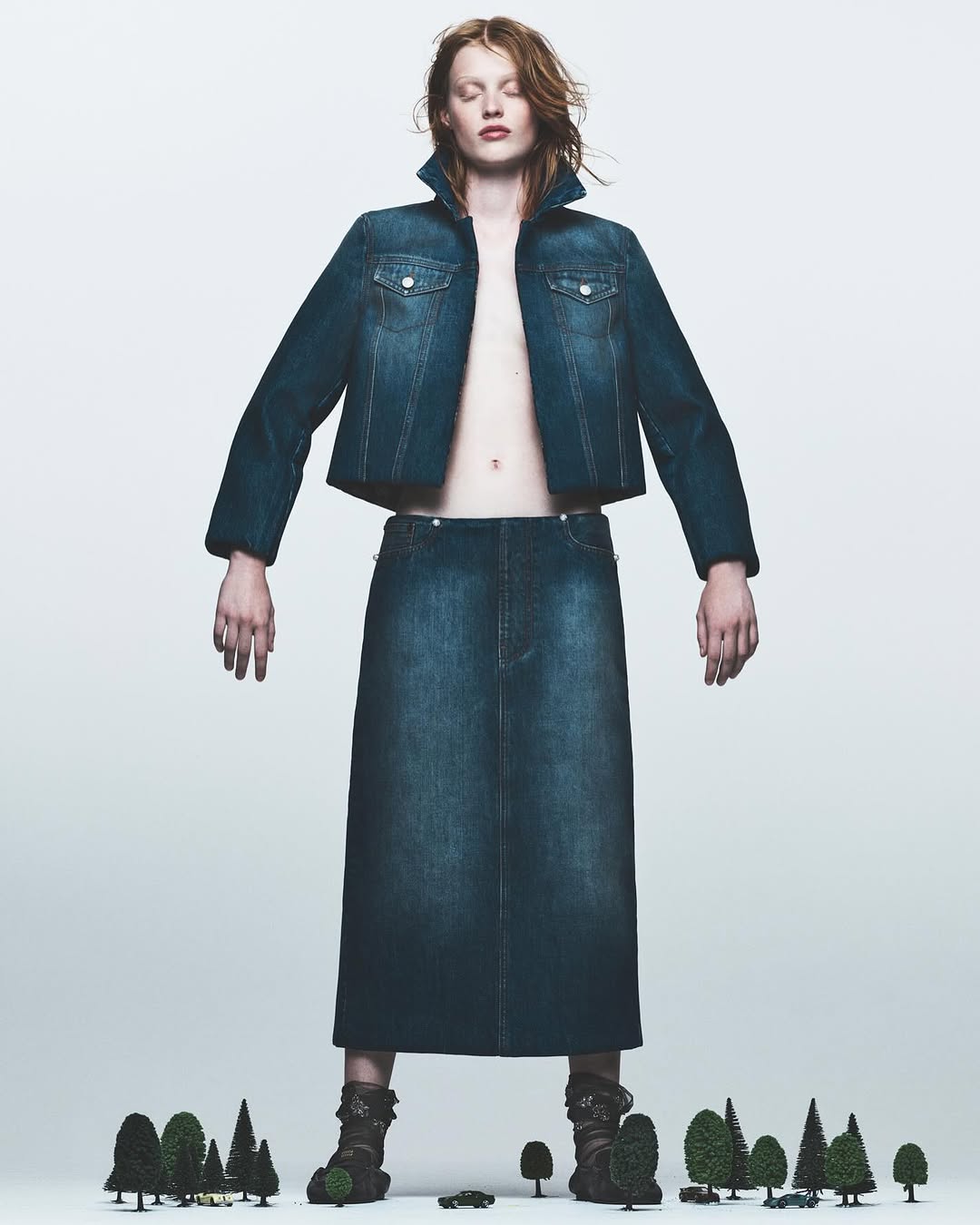 A woman in a denim jacket and skirt poses in front of a white background with miniature trees, in a fashion editorial style photograph.
