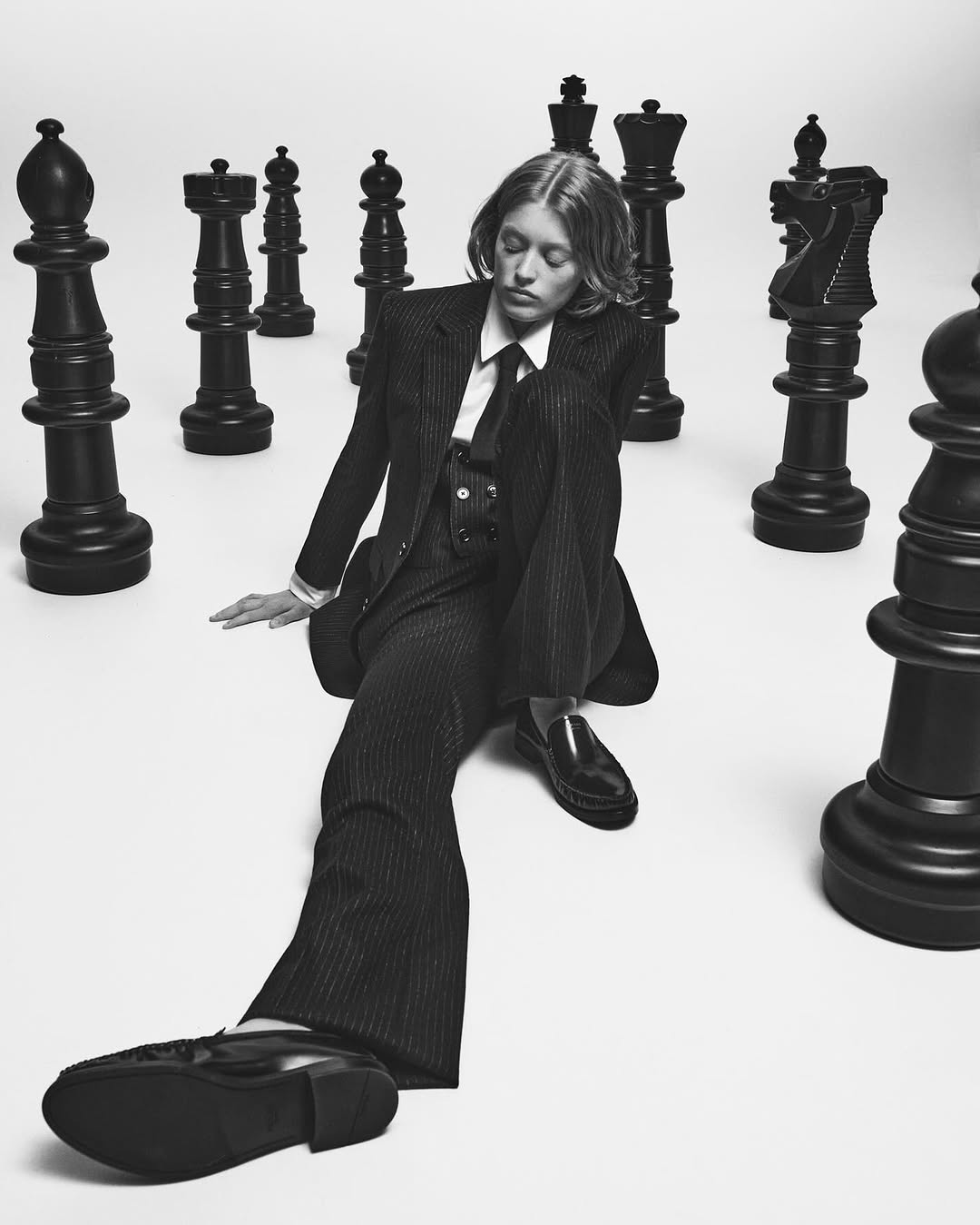 Young woman in pinstripe suit poses seated among giant chess pieces in a studio, looking down, against a white background, monochrome aesthetic.