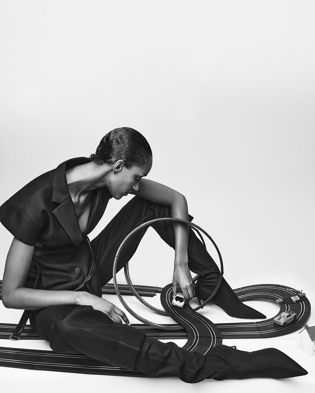 Adult female in a dark suit poses in a photo studio with a toy race track, creating a striking black and white editorial fashion image.