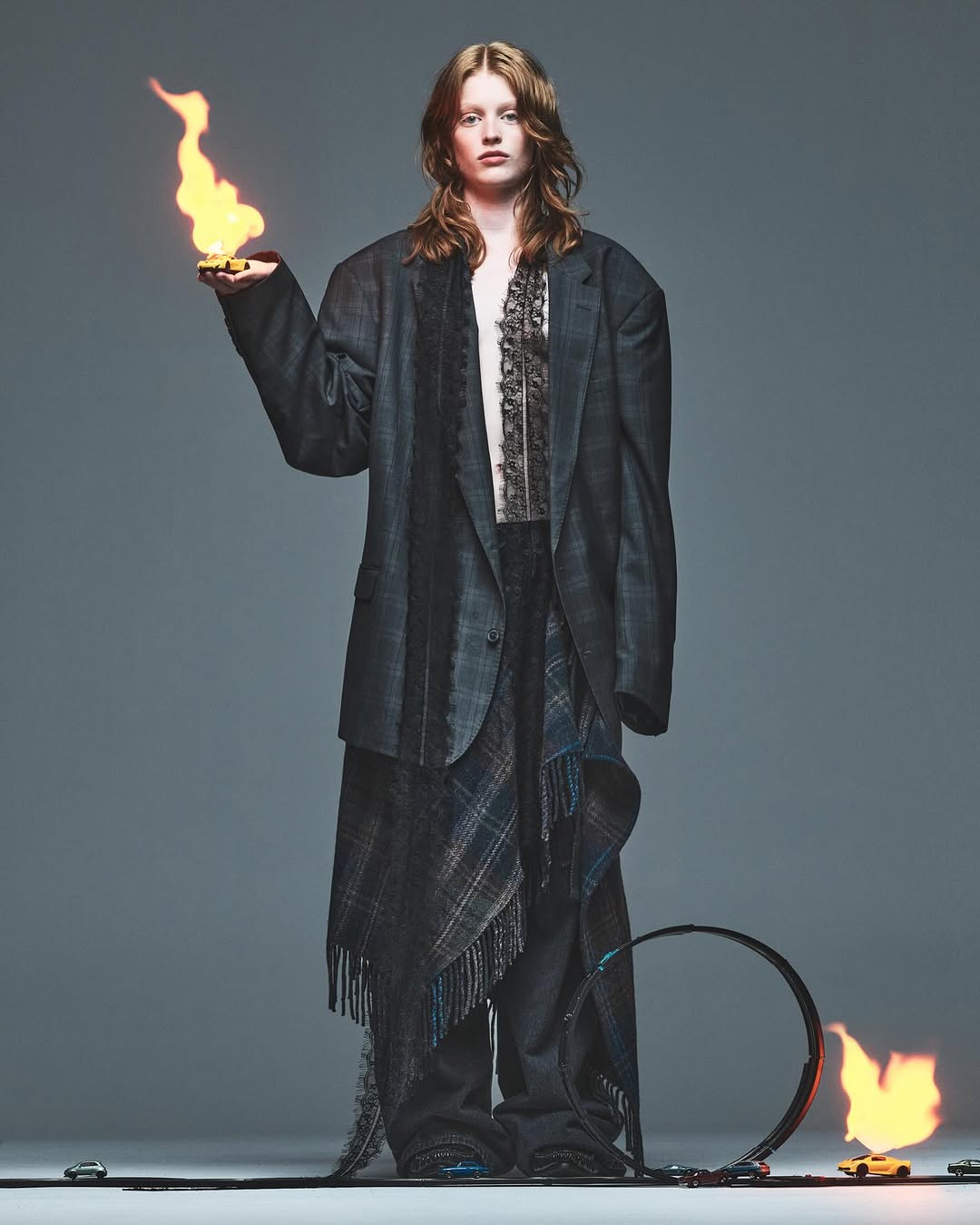 A full-length studio shot of a model wearing an oversized blazer, draped skirt, and holding flames, set against a grey backdrop with car track and loops.
