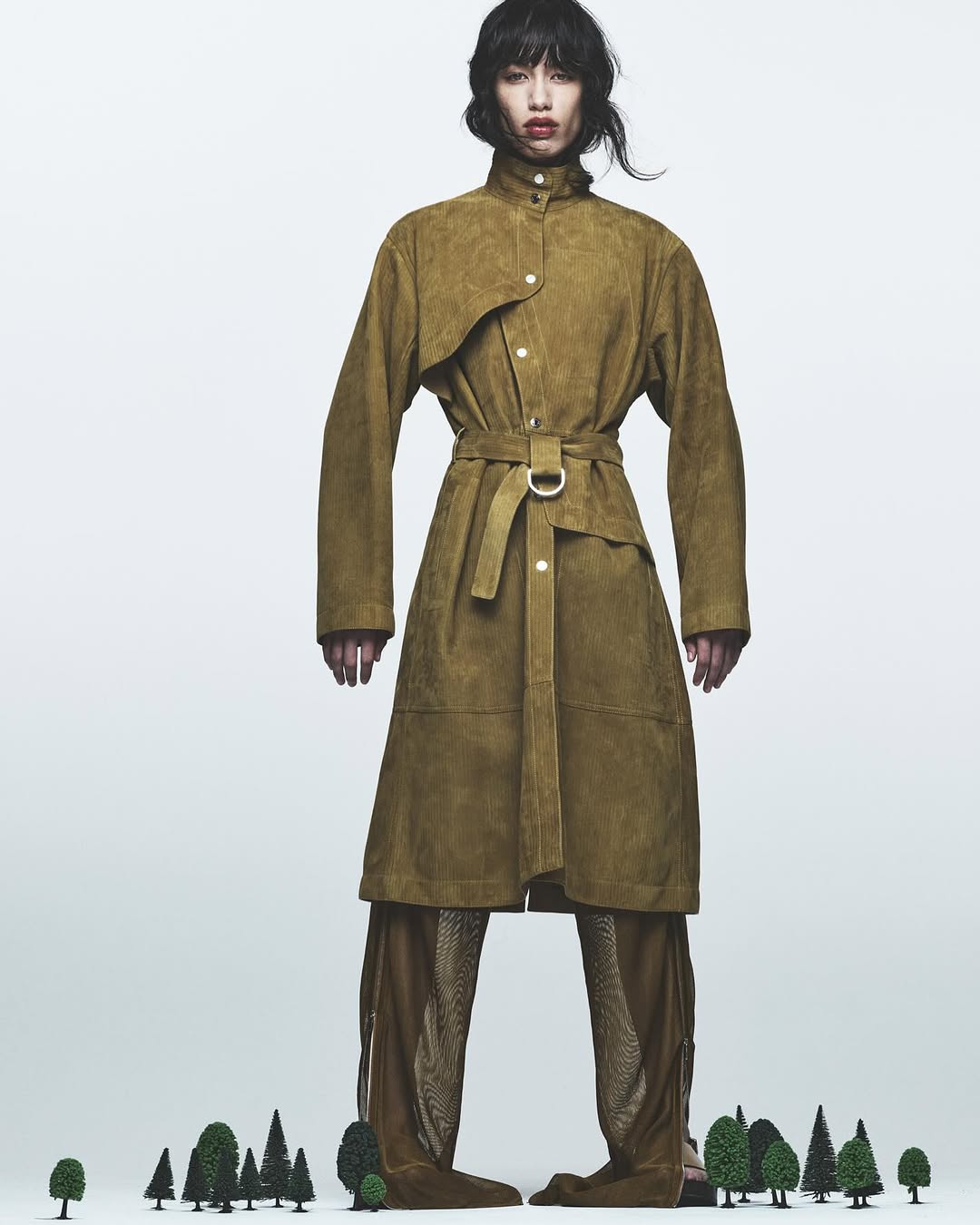 Fashion editorial shot of a slender model in a belted suede coat and see-through pants, standing against a white backdrop.