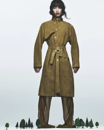 Fashion editorial shot of a slender model in a belted suede coat and see-through pants, standing against a white backdrop.