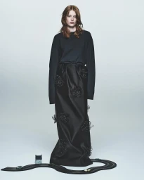 Woman in dark long-sleeve top and black textured skirt standing over a toy race track against a grey background