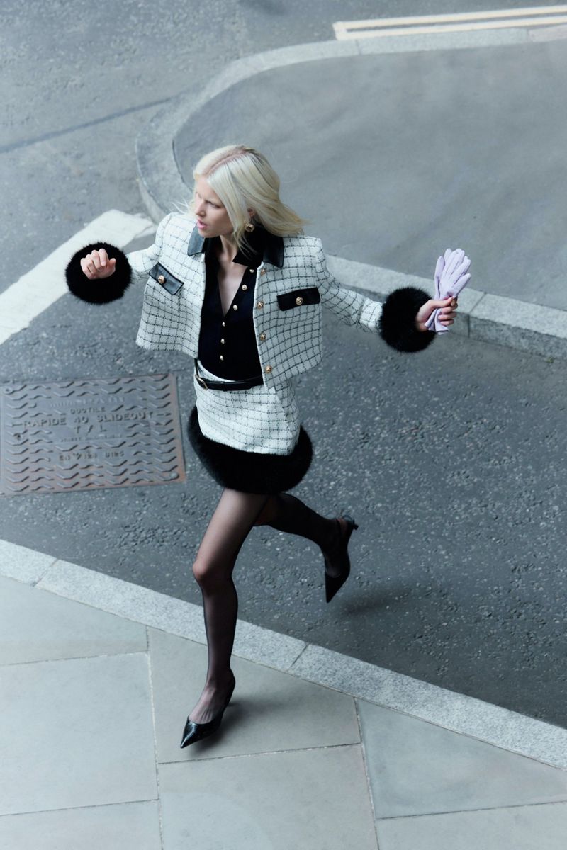 Blonde woman in a checkered white and black skirt suit and black tights runs on a grey sidewalk with pastel gloves in hand.