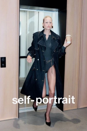 Fashionable woman in a trench coat exits an elevator holding a coffee cup, wearing heels and a bodysuit with jewelry details.