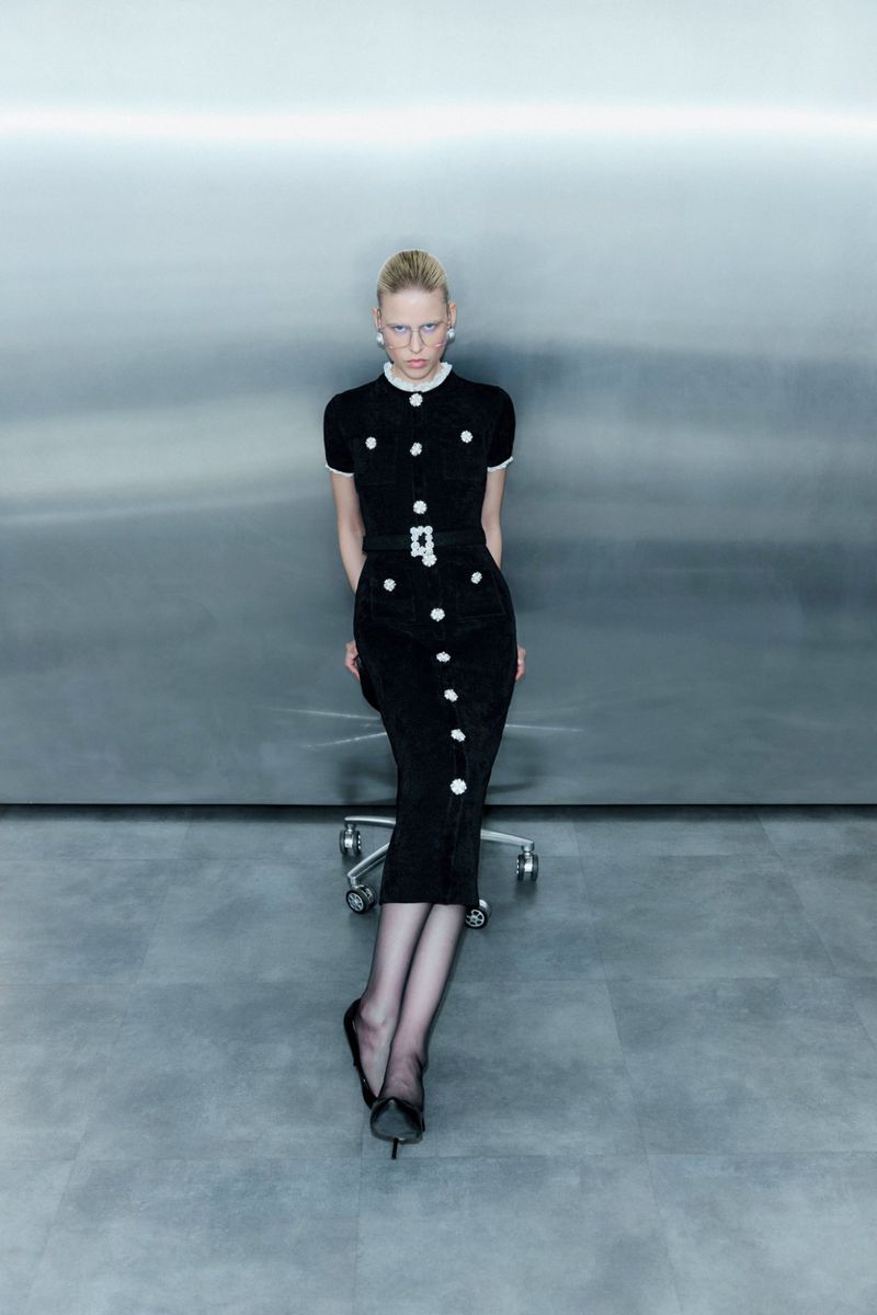 Adult female in a black dress with white buttons poses sitting on a rolling chair in a studio with gray wall and floor.