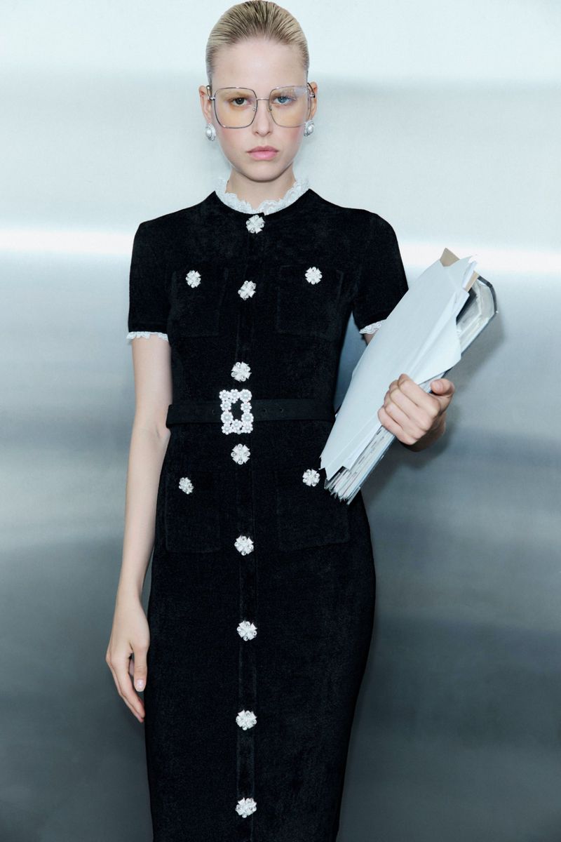 Woman in a black dress with white floral embellishments, holding papers, wears translucent eyeglasses against a gray studio backdrop.