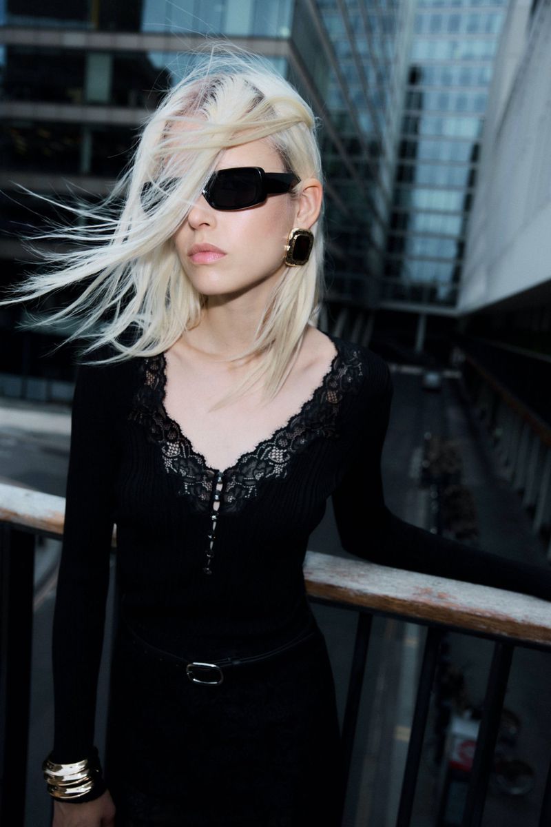 Woman with platinum blonde hair poses in an urban setting wearing sunglasses, earrings, long-sleeved dress, belt, and metal bracelets.