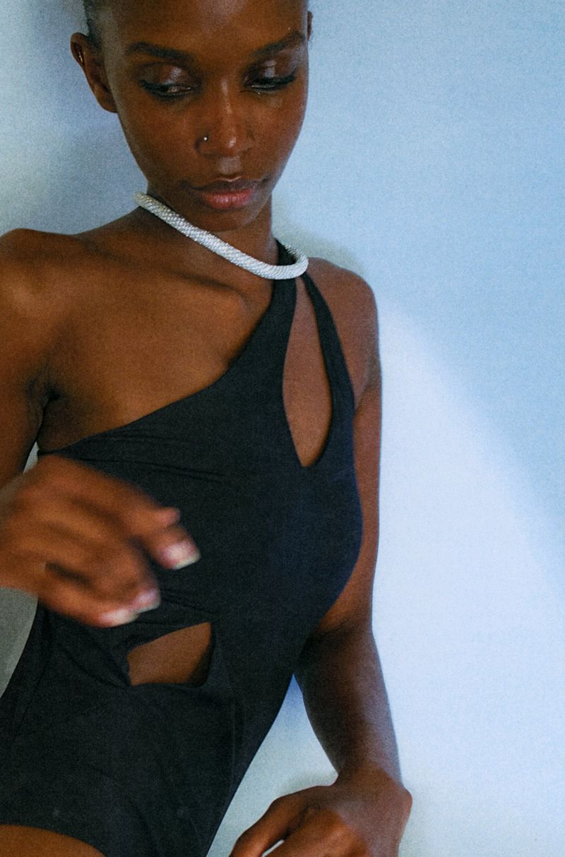 Model Ornella Umutoni wearing a black cutout one-shoulder swimsuit and a white beaded choker against a pale background.