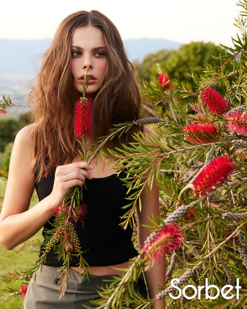 A young woman with long brown hair poses in an outdoor setting with red bottlebrush flowers and green foliage against a soft hillside backdrop.
