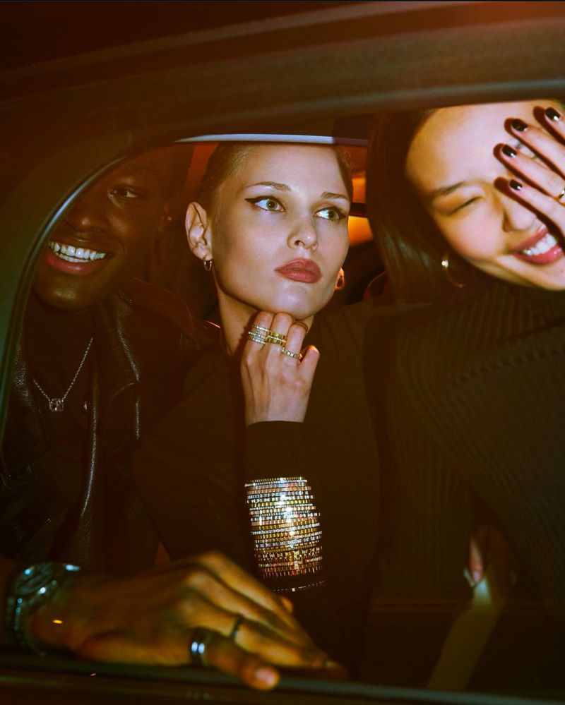 Three women in a car, smiling and wearing jewelry. The woman in the center is the main focus, with dark clothing and bracelets.