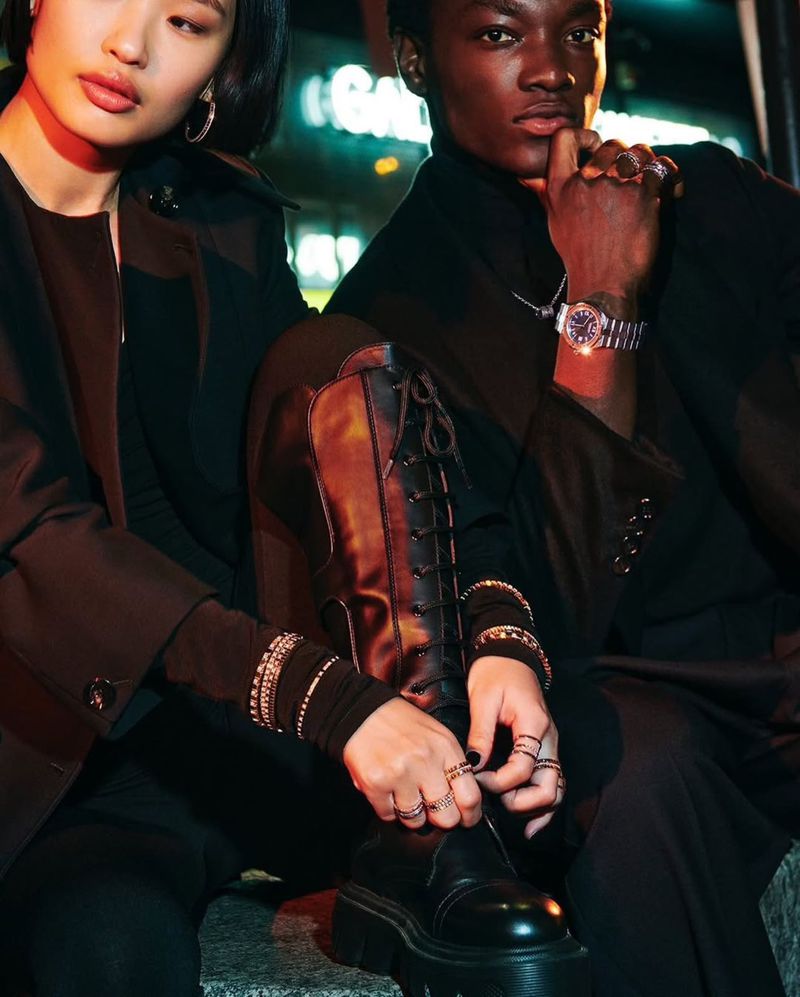 Two fashion models in black outfits pose outdoors at night, one putting on a lace-up boot, showcasing watches and rings.
