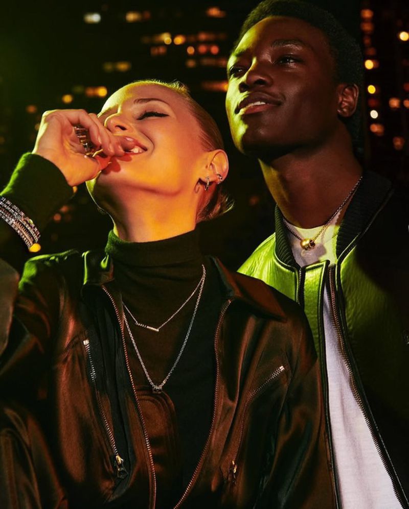 Two people pose in a medium shot at night with city lights blurred in the background. A blonde woman puts food in her mouth.
