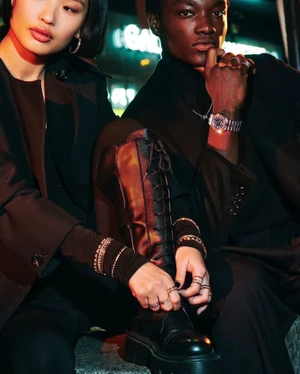 East Asian woman tying a black lace-up boot beside a Black man wearing a luxury wristwatch and dark coat at night.