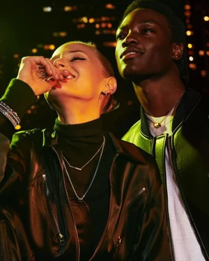 Two models wearing layered jewellery and jackets outdoors at night with warm city lights in the background.