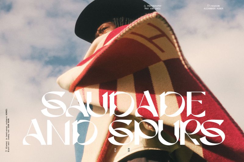 Editorial fashion shot featuring a person in a cowboy hat draped in a striped Hermes blanket standing against a partly cloudy sky.