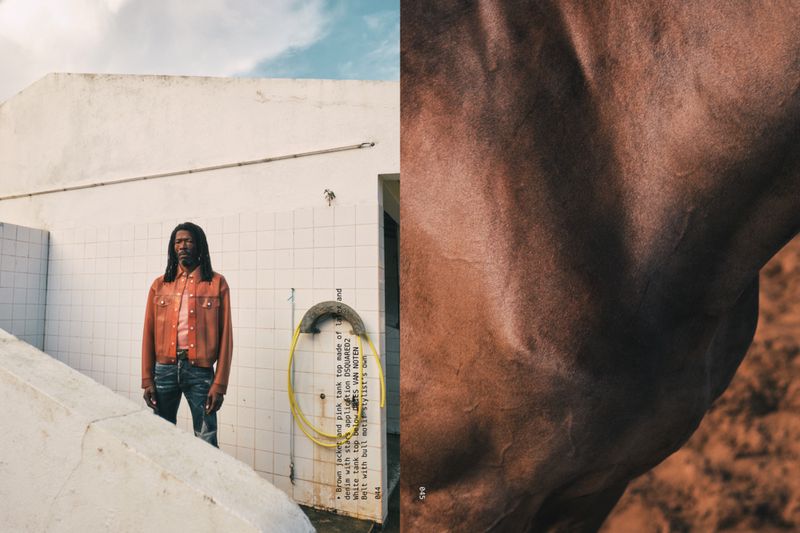 An adult male with long dreadlocks wears an orange denim jacket and dark jeans, standing outside a white-tiled building with brown horse hair nearby.