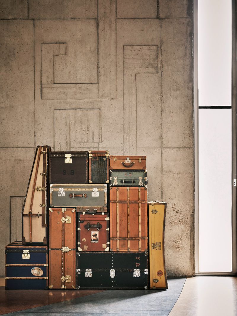 Stack of vintage Louis Vuitton trunks and suitcases against a textured concrete wall, next to a bright window, evoking a sense of travel and luxury.