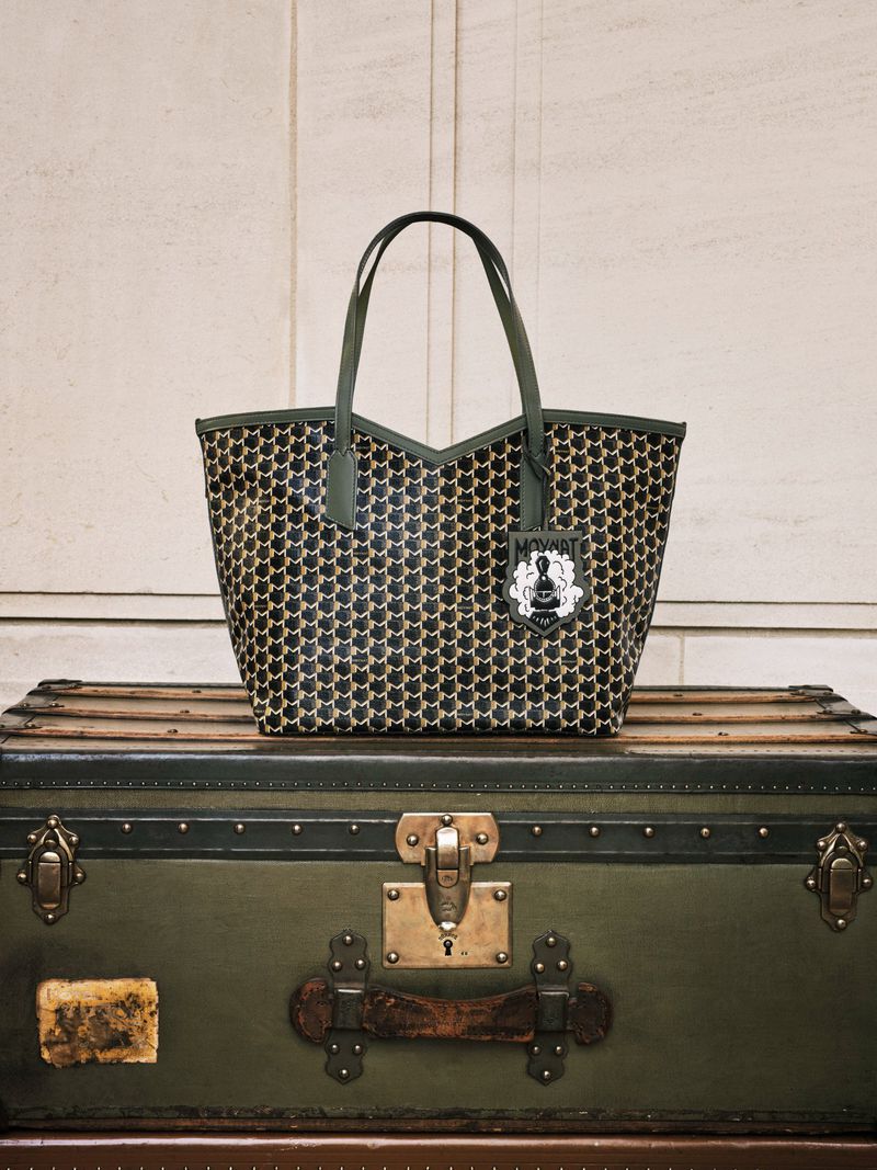 Patterned tote bag with green handles and trim, resting on an olive-green vintage trunk with brass hardware and a neutral background.