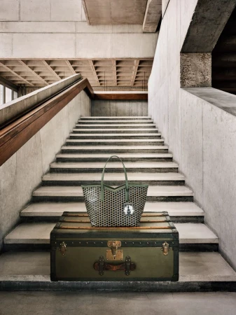 Olive green tote bag with a geometric pattern resting on an antique trunk set against a backdrop of concrete stairs and walls.