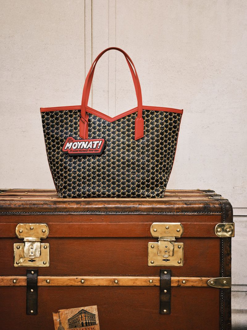 Black and tan patterned tote bag with red trim and handles and the 'Moynat!' logo, sitting on top of a brown antique steamer trunk.