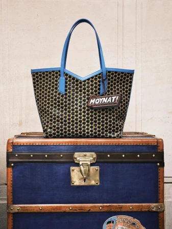 Patterned tote bag with blue trim sits atop a vintage-style blue trunk with brass hardware, set against a cream-colored wall.
