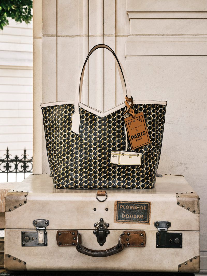 A patterned tote bag with cream-colored handles sits atop a vintage suitcase against a neutral stone background indoors.