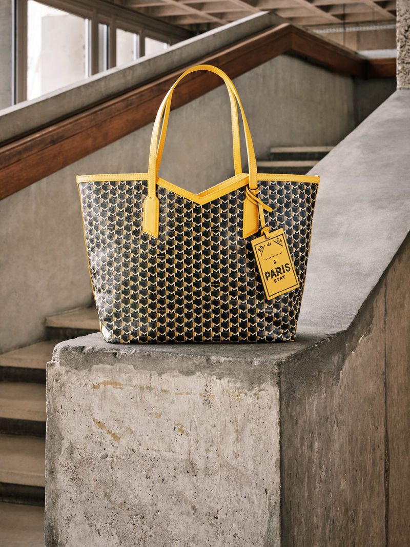 Patterned tote bag with yellow trim and handles resting on concrete steps indoors, featuring a hanging tag.