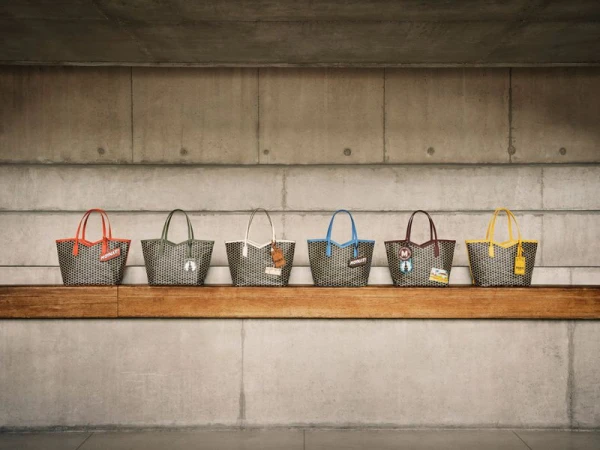 Six grey patterned tote bags with colorful handles sit in a row on a wooden bench against a concrete wall in an indoor setting.