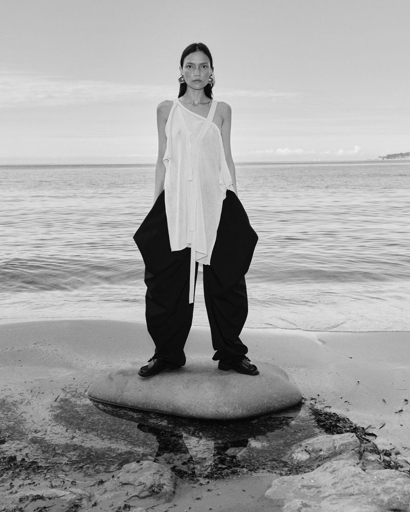 Charlotte Carey standing on a flat stone on a sandy beach wearing an asymmetric white tunic and wide black trousers.