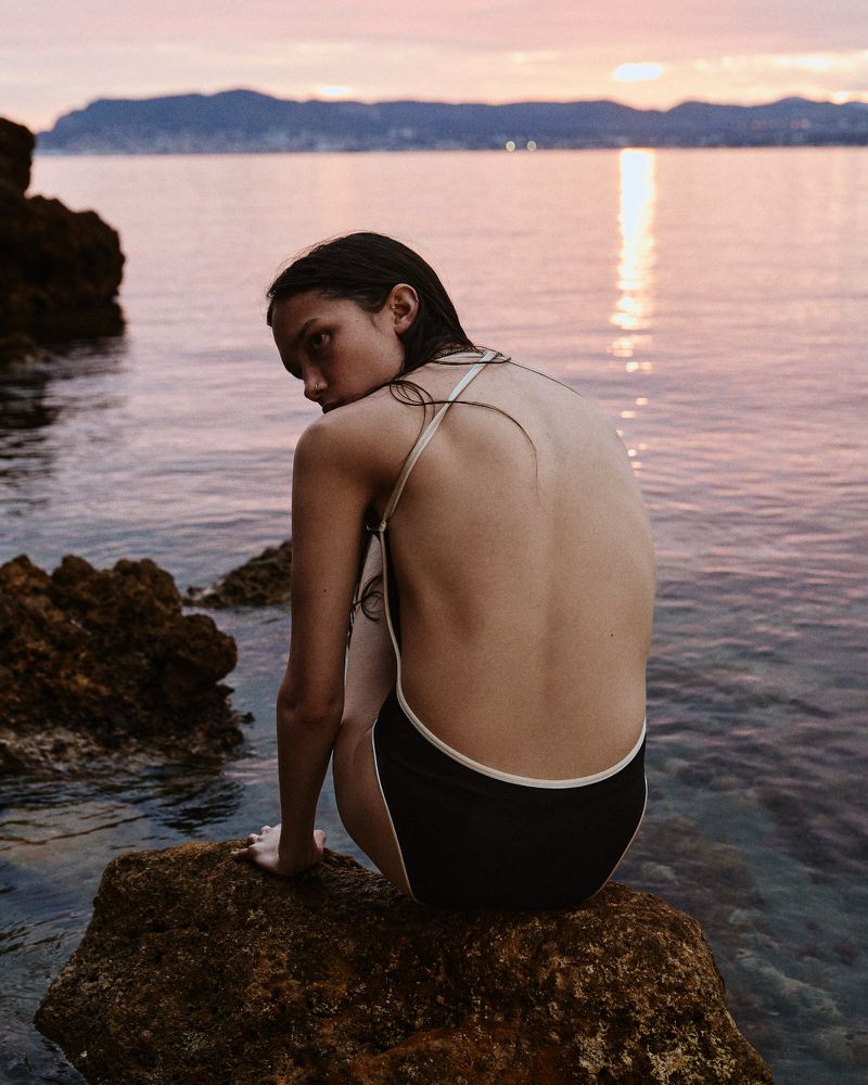 A young woman in a swimsuit sitting on rocks, looking over her shoulder with an ocean and sunset backdrop, creating a serene outdoor fashion shot.