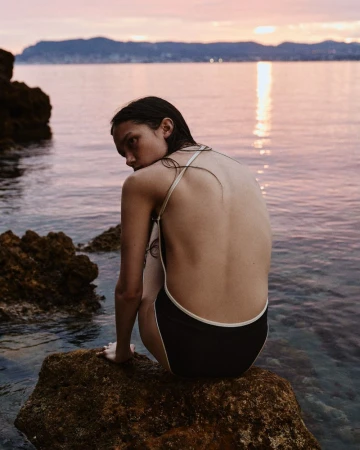 A young woman in a swimsuit sitting on rocks, looking over her shoulder with an ocean and sunset backdrop, creating a serene outdoor fashion shot.