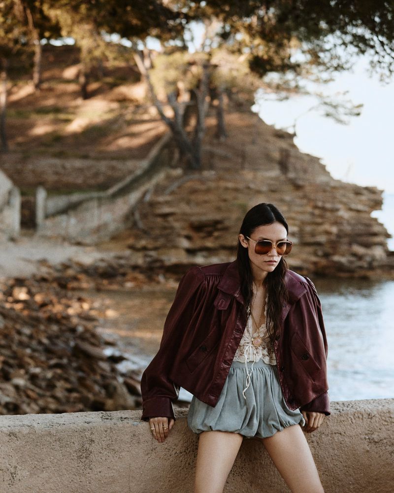 A fashionable adult woman with sunglasses poses outdoors in a burgundy jacket and pale-blue shorts near a stone wall and rocky coastline.