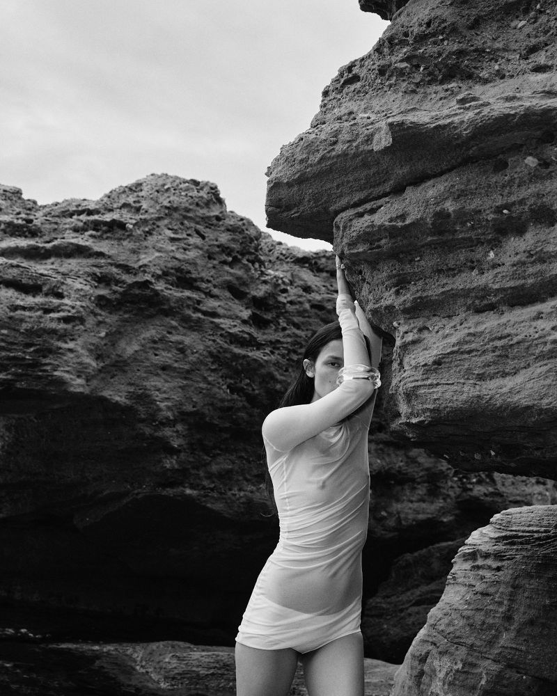 Monochrome portrait of a woman in a sheer dress posing among rock formations with neutral expression and hands up, outdoors.