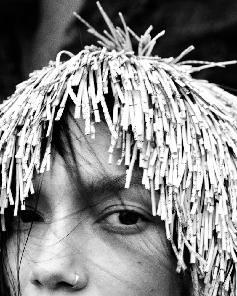 Close-up black and white portrait of a person with dark hair and a nose ring wearing a hat made of shredded paper strips.