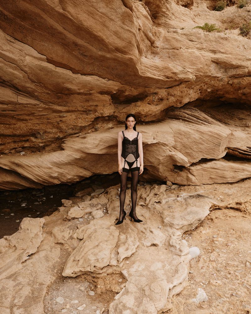 A woman in lingerie stands on rocks against a layered-rock background, highlighting fashion in a natural setting.