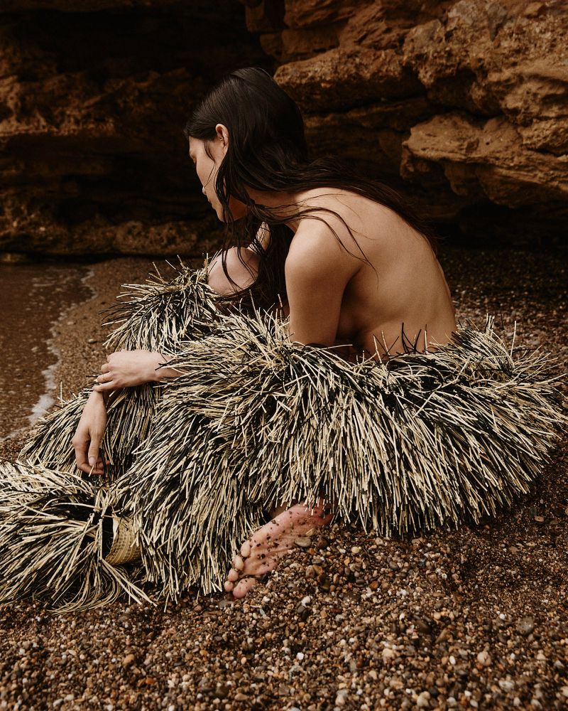 A nude woman with long hair sits on a gravelly beach wearing a spiky straw-like skirt near rocks and water, viewed in a medium shot.