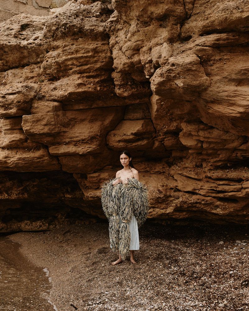 Woman on a beach with a rock formation. She is wearing a fringed garment over a white dress. The scene has a natural, earthy tone.
