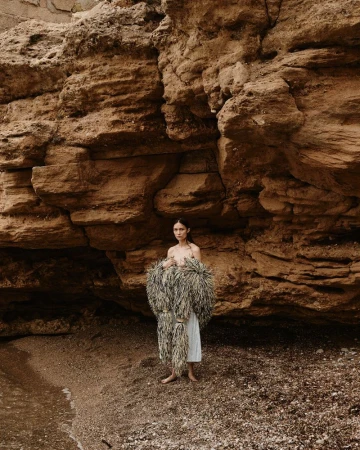 Woman on a beach with a rock formation. She is wearing a fringed garment over a white dress. The scene has a natural, earthy tone.