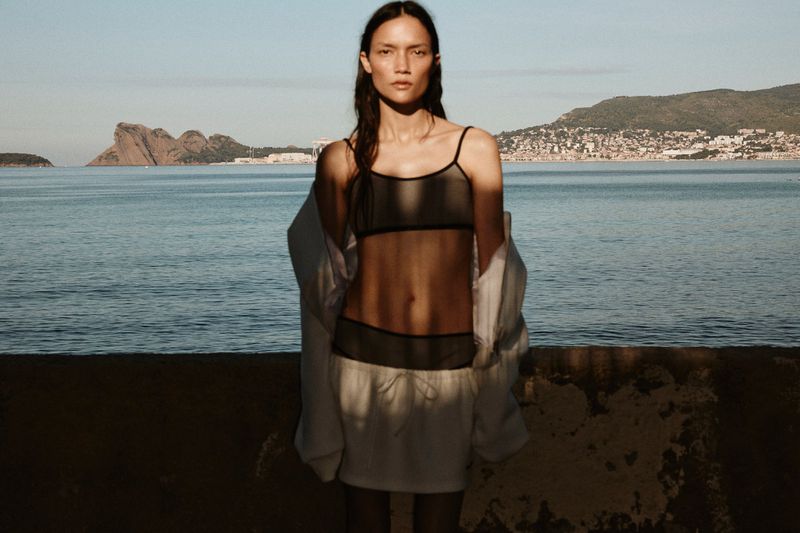 Adult woman in black bra and white shorts posing near the sea with a city in the background during daytime. Fashion shot.