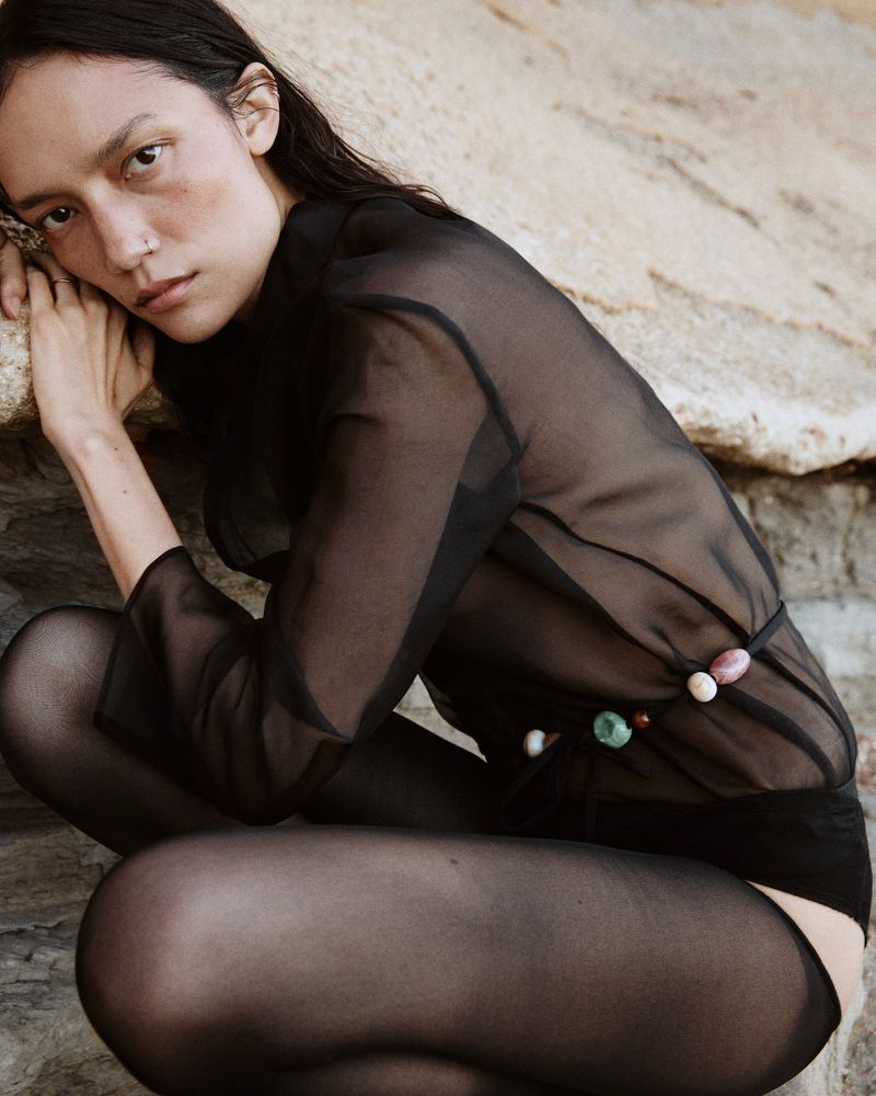 Portrait of a slender woman with dark hair, wearing a sheer top and tights, crouching near rocks in an outdoor environment.