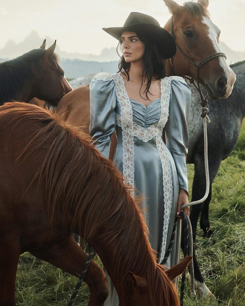 Cowgirl in a satin dress surrounded by brown horses in an open field at sunset. Woman poses in an outdoor fashion editorial.