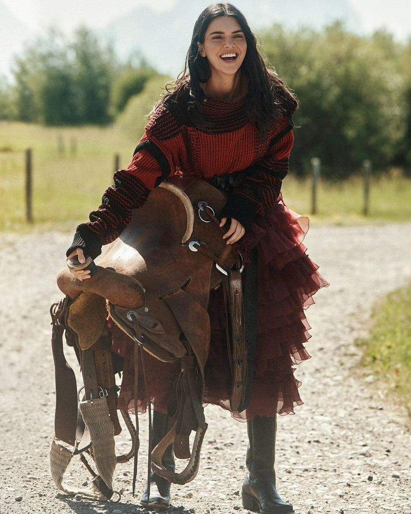 Smiling woman poses with horse saddle on rural road. The model is wearing a maroon knit sweater, layered skirt and boots in a natural setting.