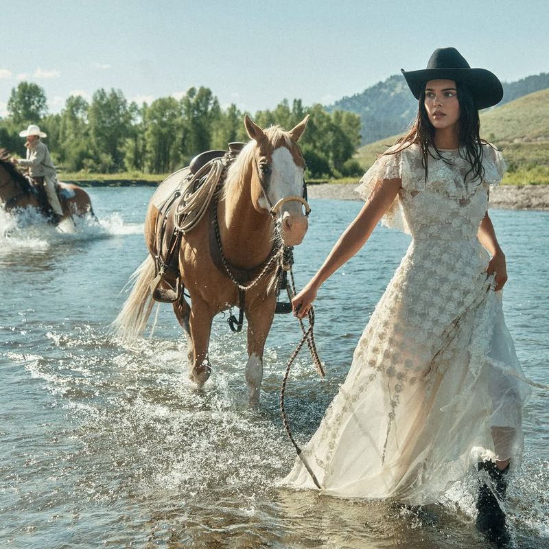 A woman in an elegant dress leads a horse through a shallow river, with another rider in the background, set against a mountain backdrop.