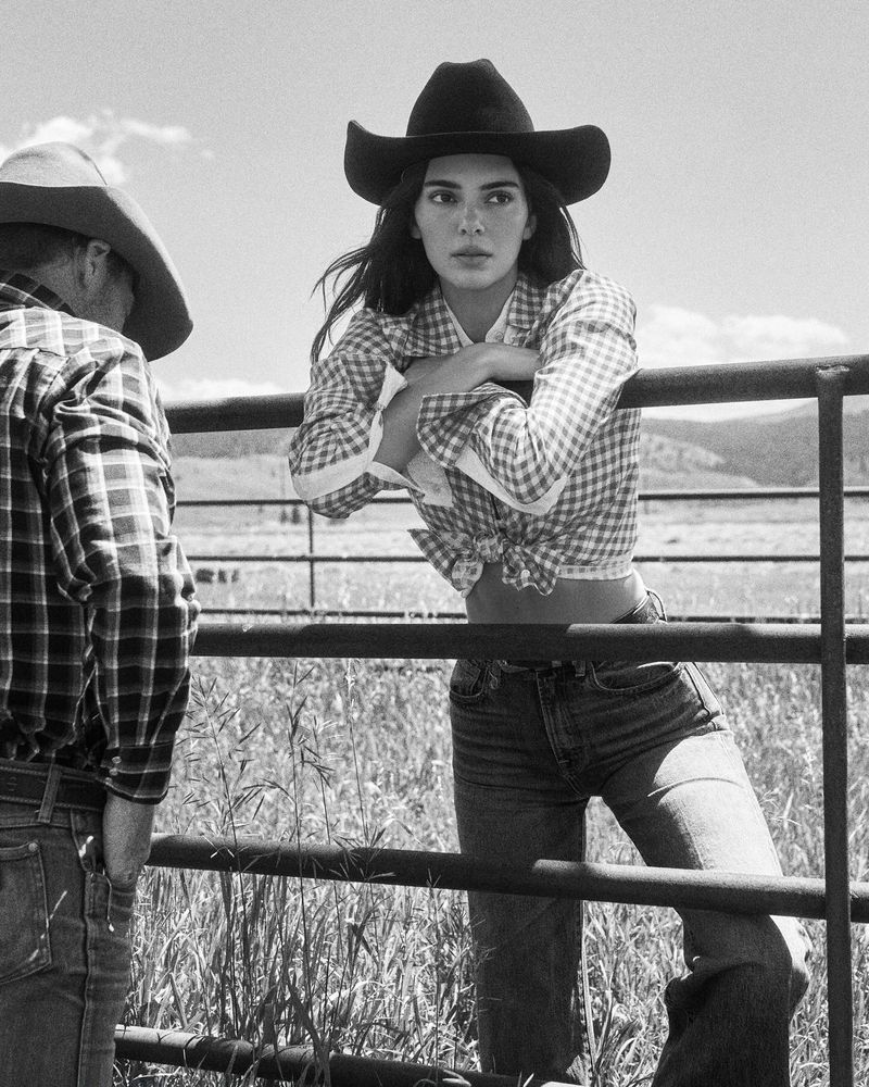 Black and white shot of a woman in a cowboy hat leaning on a fence with another person wearing a hat standing in the background.