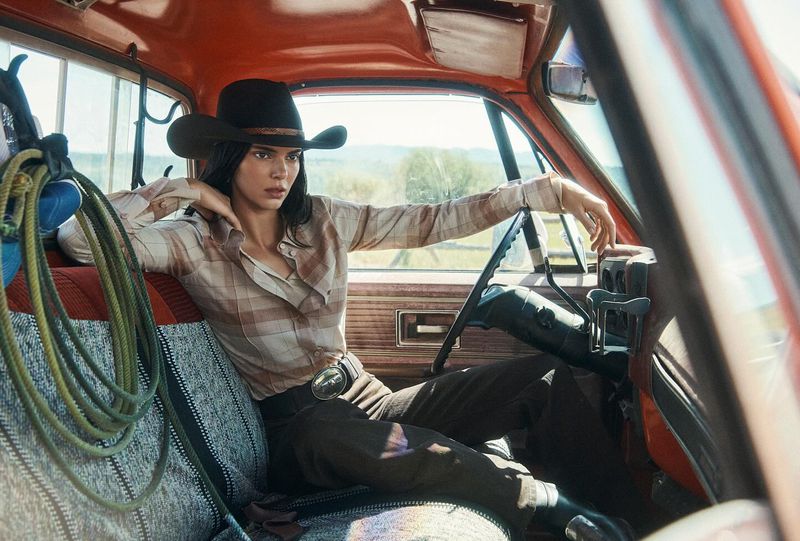 A fashionable young woman in a cowboy hat sits inside a vintage red truck, wearing a Western-style outfit, with rural scenery in the background.