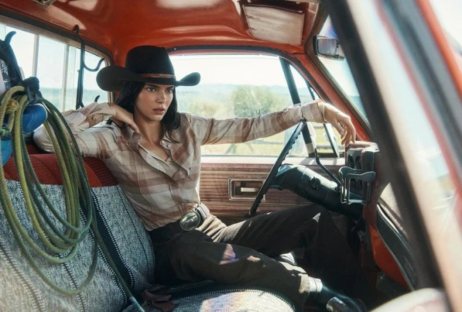 A fashionable young woman in a cowboy hat sits inside a vintage red truck, wearing a Western-style outfit, with rural scenery in the background.