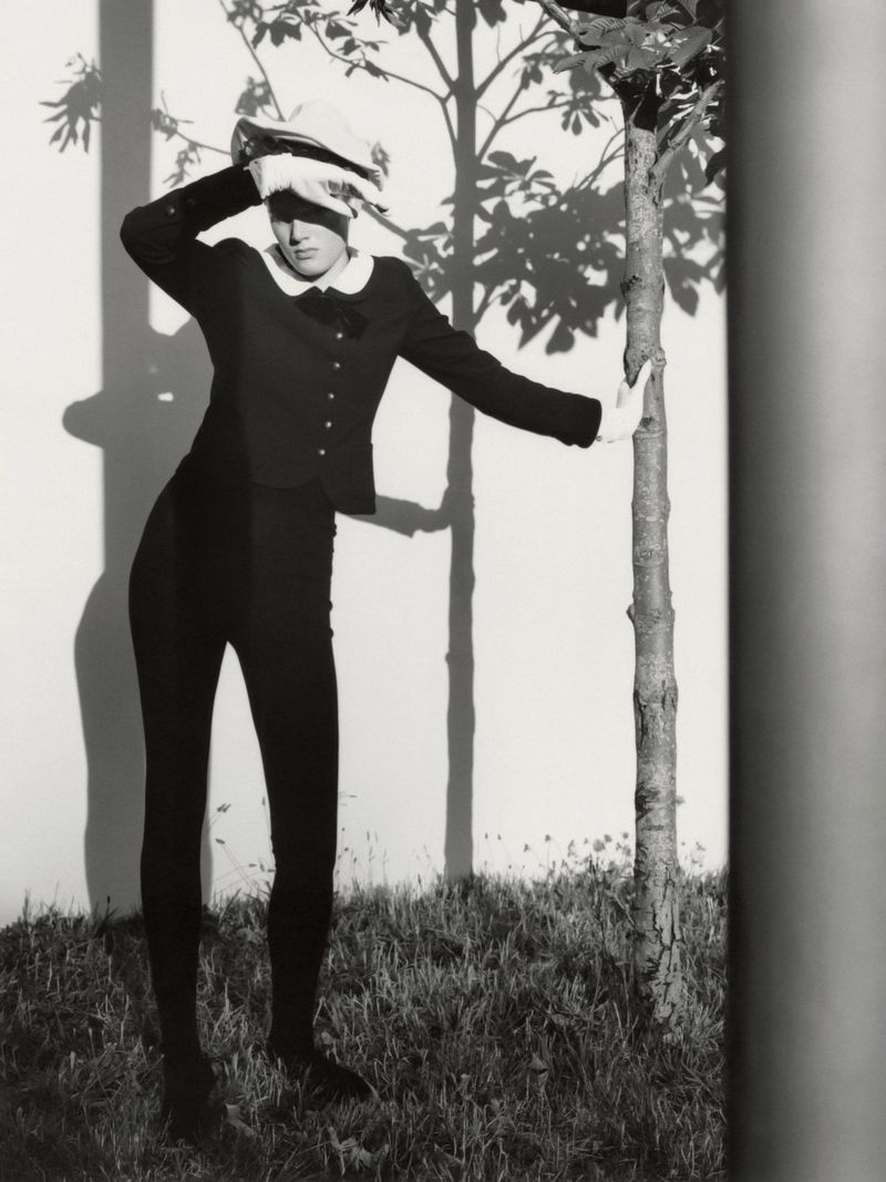 Model Noor Khan wearing a dark buttoned jacket with a white collar, posing by a slender tree against a sunlit wall in black and white.