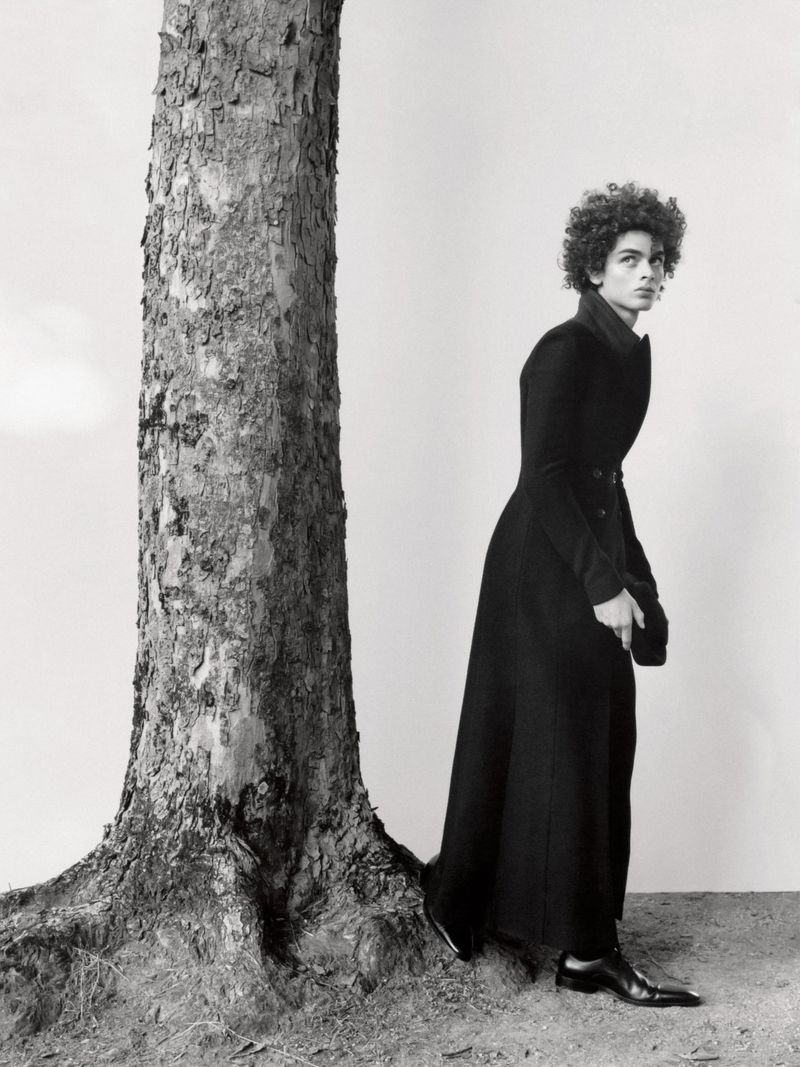 Black and white portrait of an adult with curly hair and a long dark coat posing next to a large tree trunk outdoors.