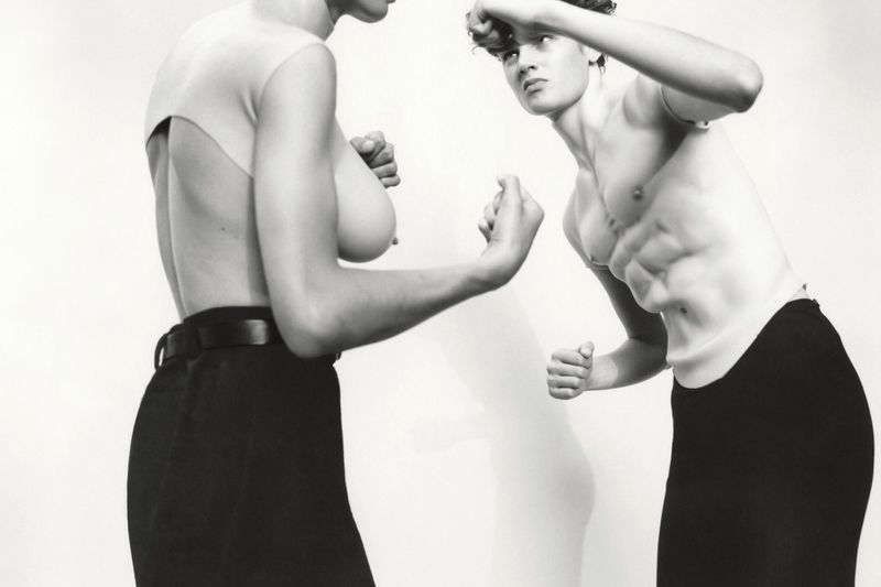 Two shirtless people face each other in a black and white studio shot, with fists raised, wearing black pants against a white background.